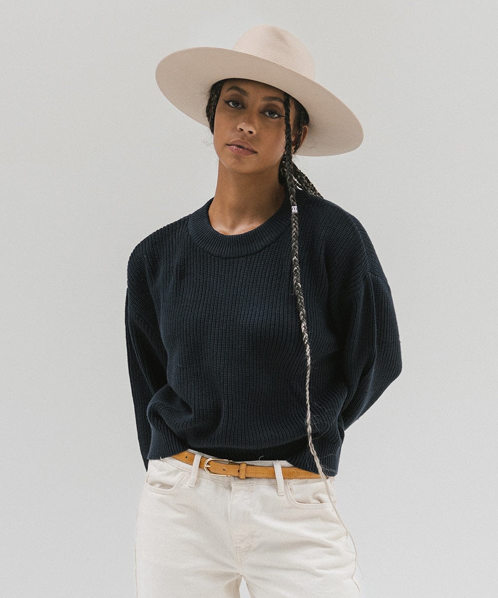 Woman wearing a cream wide brim felt hat with a triangle crown, a dark blue sweatshirt, brown belt, and white pants against a white background #color_cream