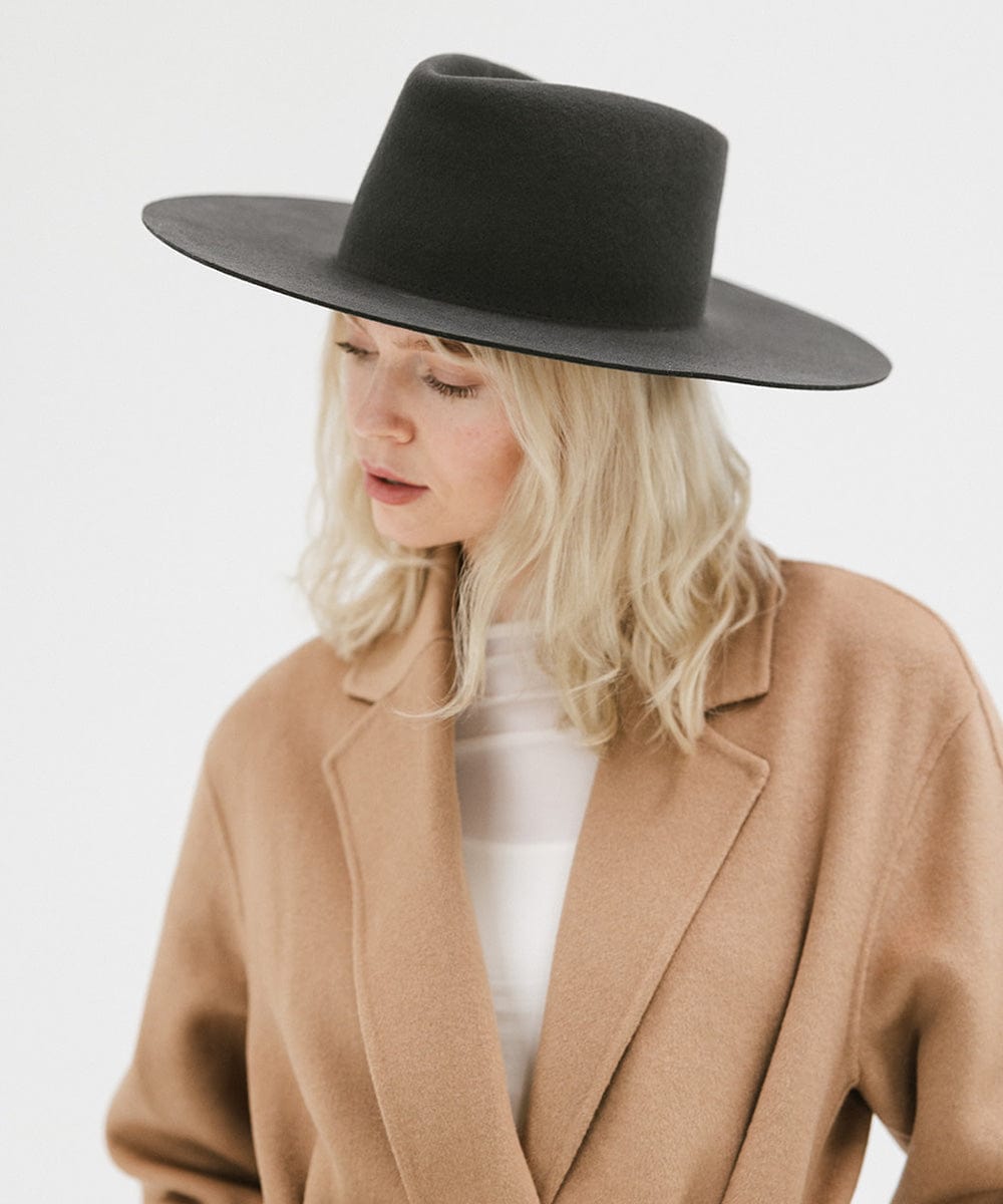 Woman wearing a dark grey wide brim felt hat with a triangle crown and a brown jacket, looking sideways against a white background #color_dark grey