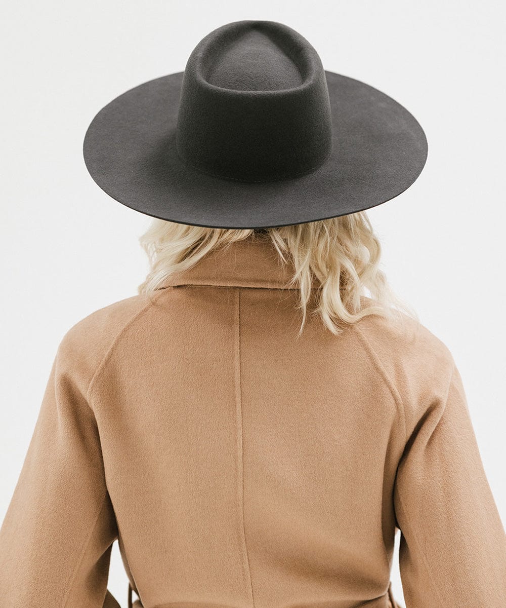 Woman wearing a dark grey wide brim felt hat with a triangle crown and a brown jacket, facing behind against a white background #color_dark grey