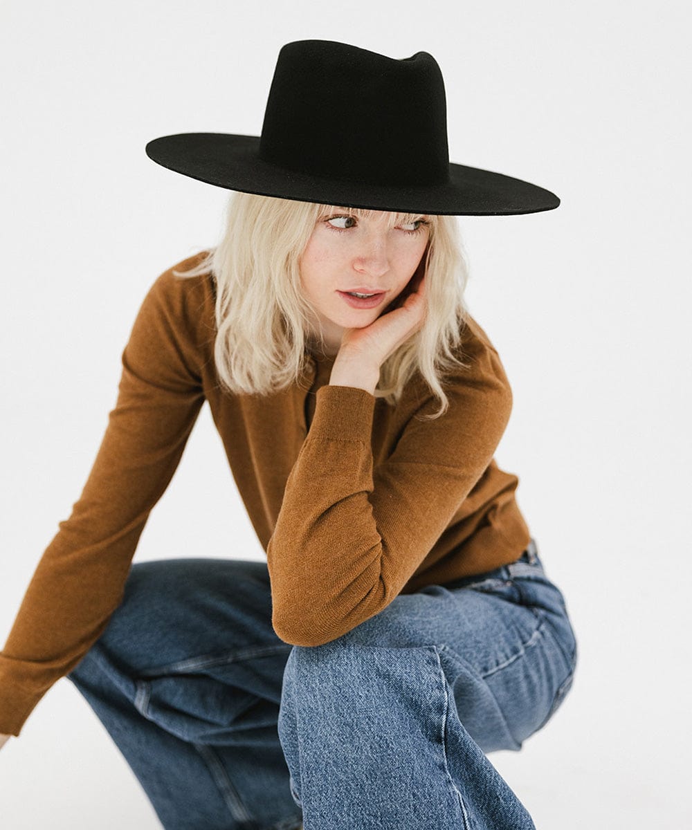 Woman wearing a black wide brim felt hat with a triangle crown, a brown shirt, and blue jeans, looking sideways against a white background #color_black