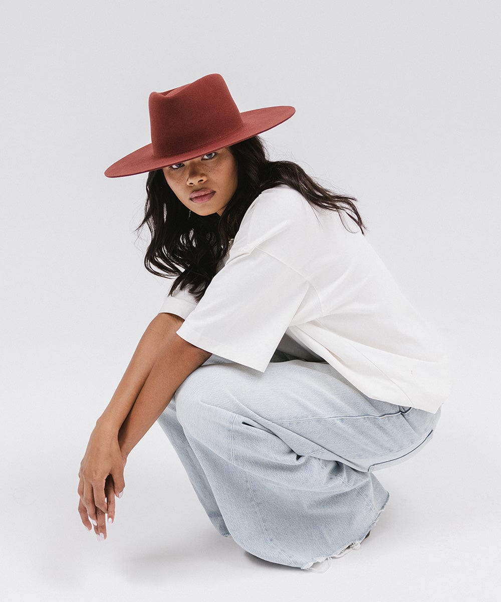 Woman wearing a rusty red wide brim felt hat with a triangle crown, a white t-shirt, and jeans, sitting against a white background #color_rusty red
