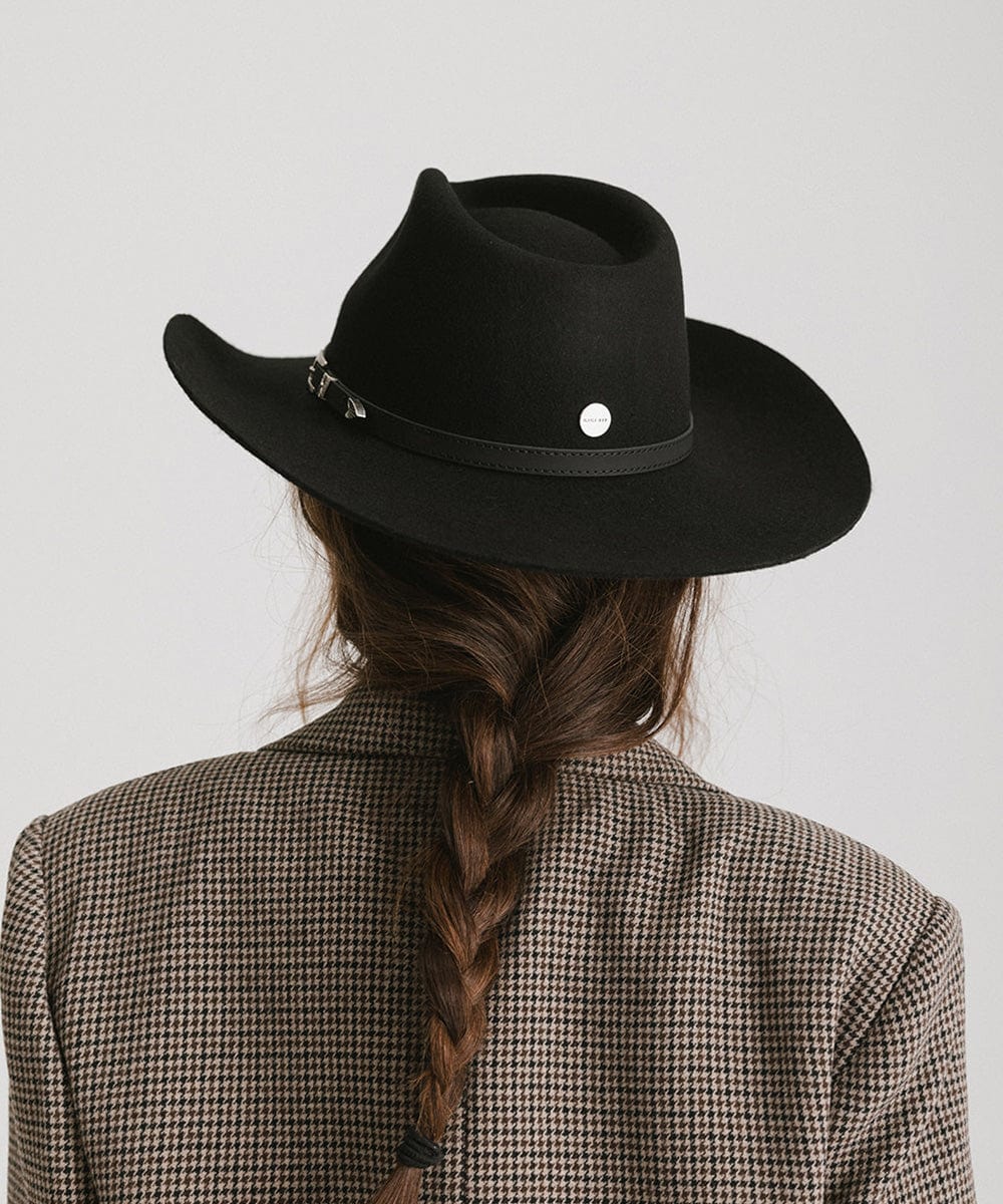Woman wearing a black cowboy hat with a hat band and a blazer, facing behind against a plain background  #color_black