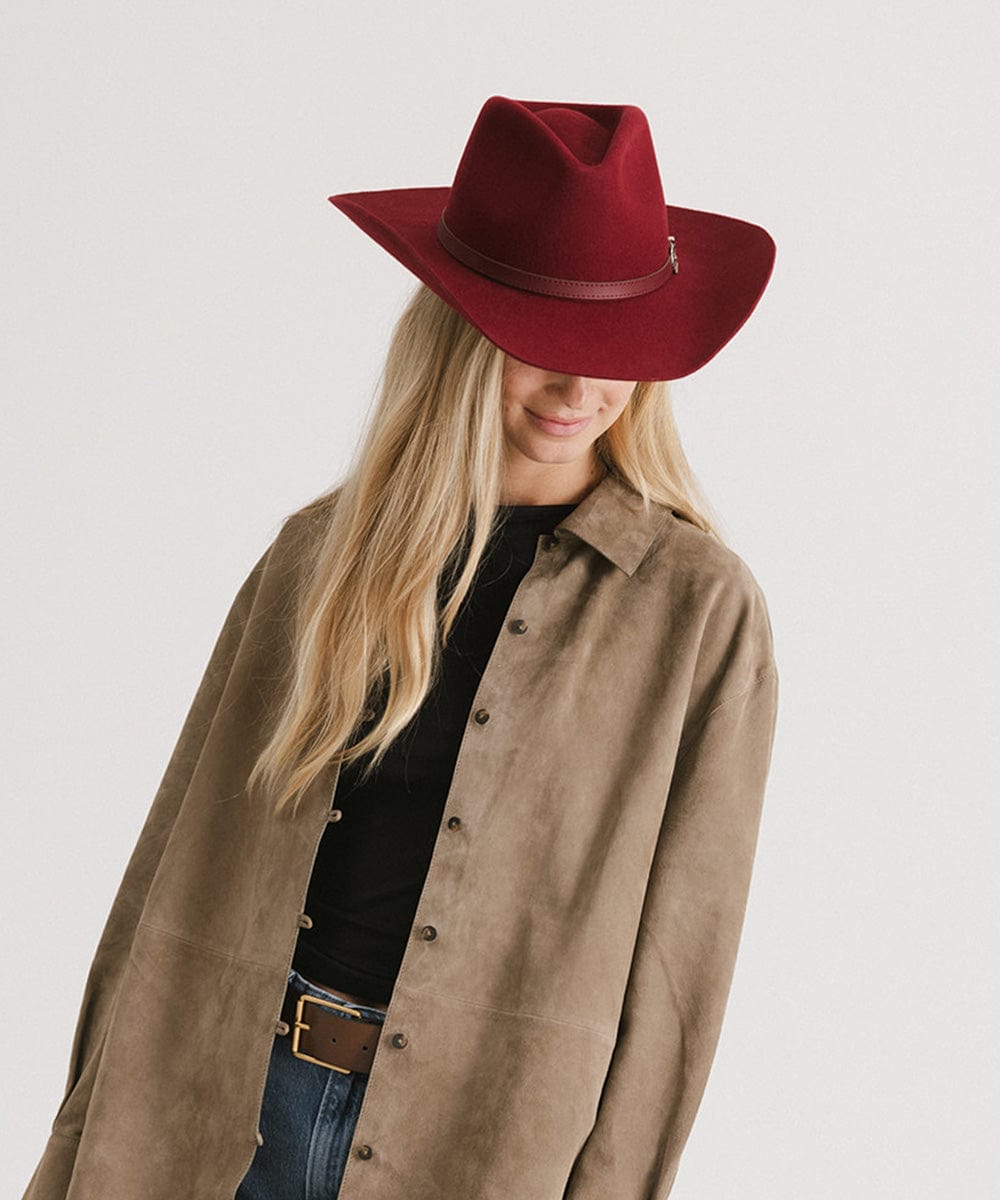 Woman wearing a burgundy cowboy hat with a hat band, a black t-shirt with a beige shirt and blue jeans against a plain background #color_burgundy