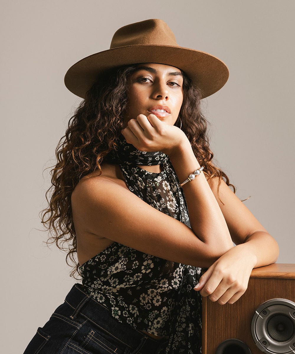 Woman wearing a caramel colored fedora hat and a floral top, leaning against a wooden speaker against a plain background #color_caramel
