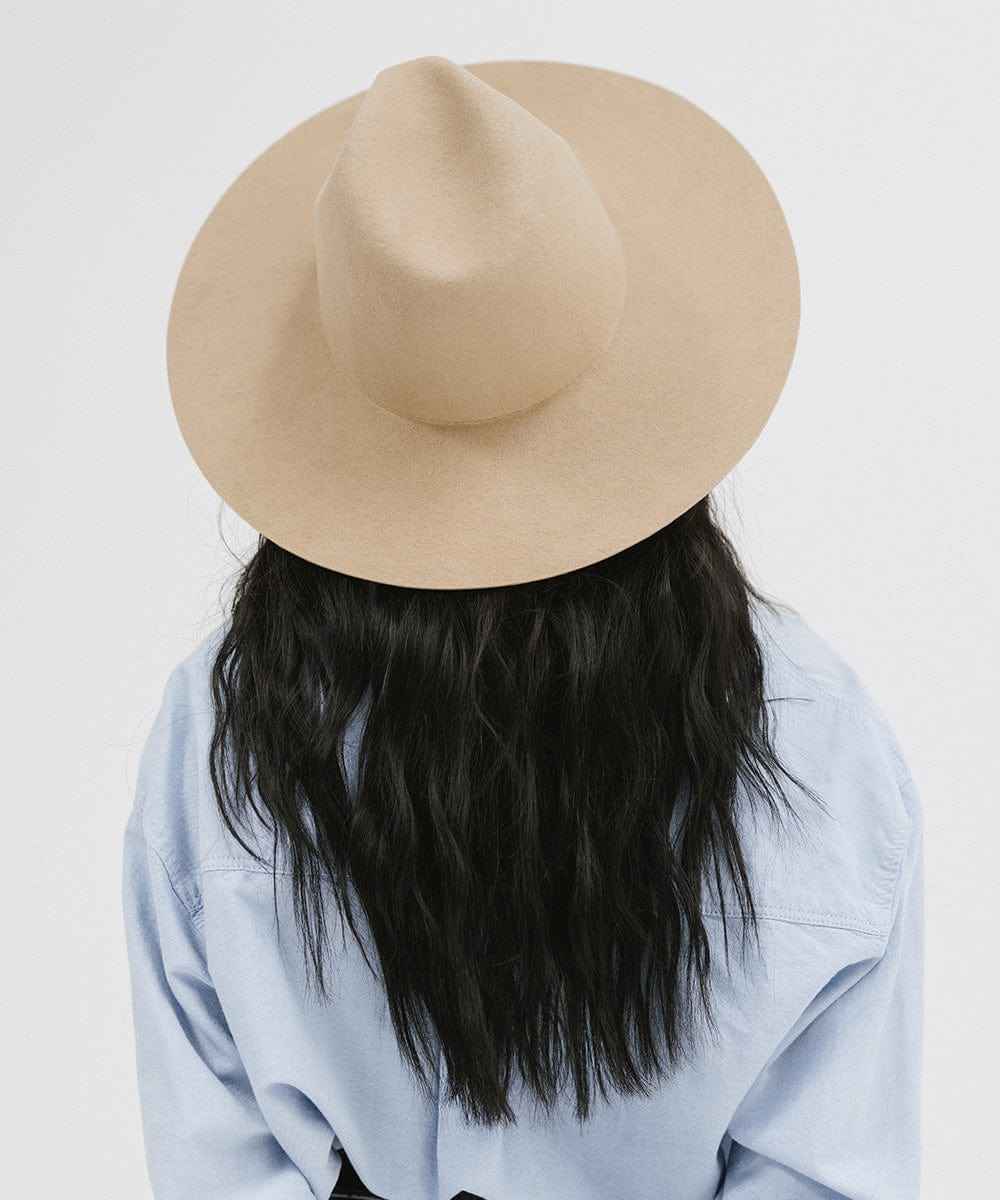 Woman wearing a tan wide brim fedora hat and a blue shirt, facing behind against a plain background #color_tan