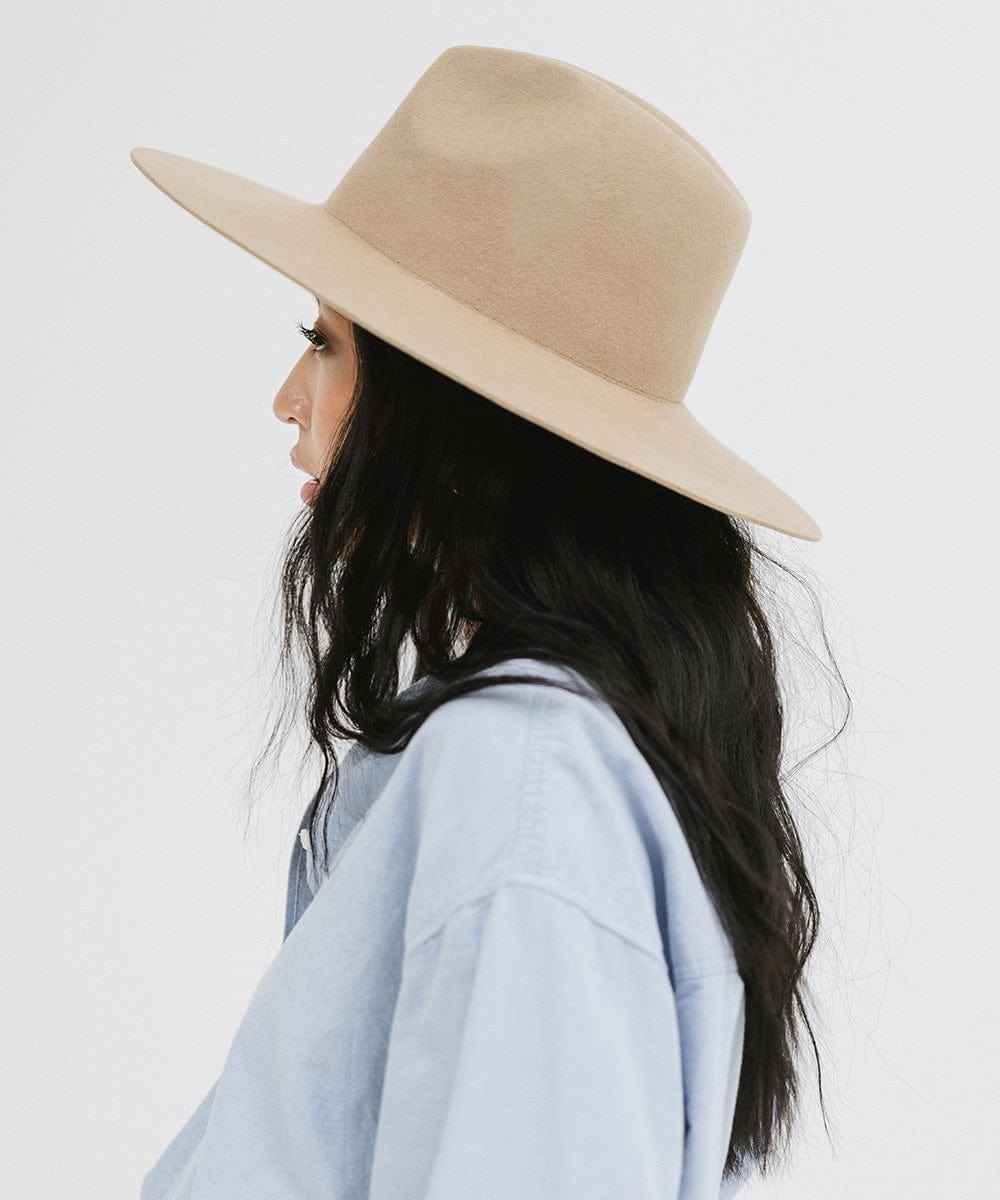 Woman wearing a tan wide brim fedora hat and a blue shirt, facing sideways against a plain background #color_tan