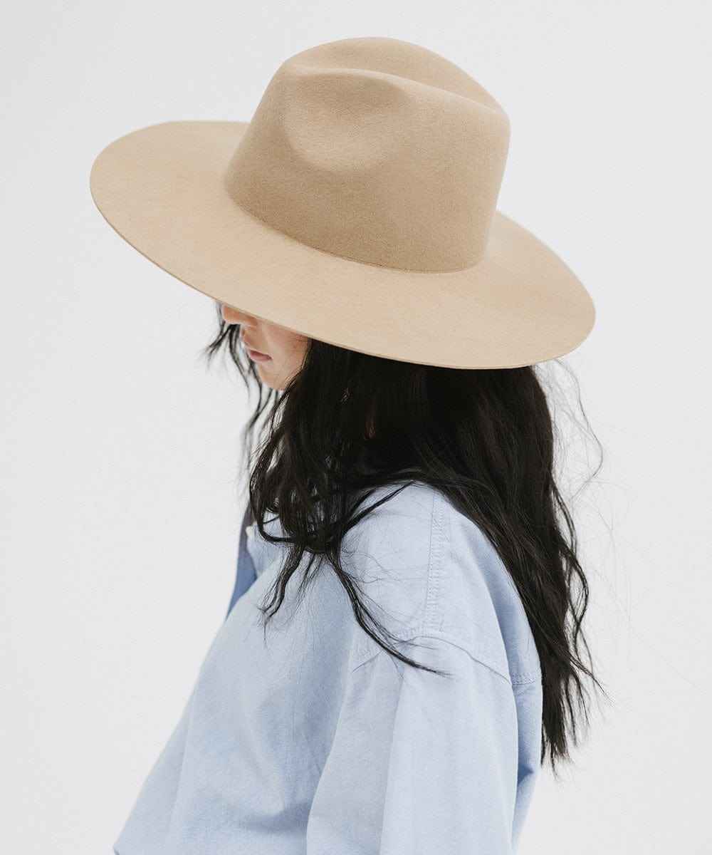 Woman wearing a tan wide brim fedora hat and a blue shirt, facing sideways against a plain background #color_tan