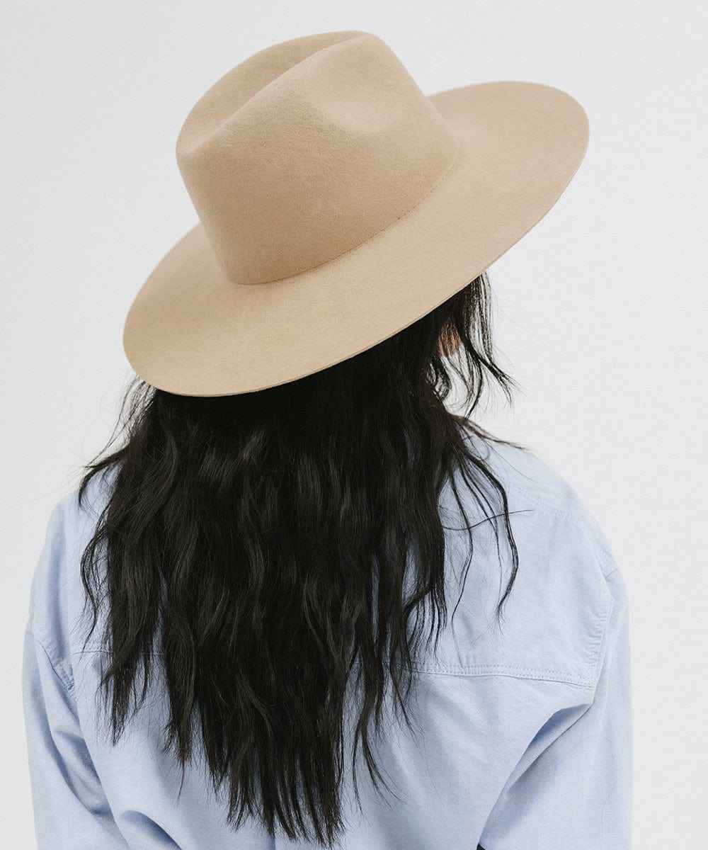 Woman with long black hair wearing a tan wide brim fedora hat and blue shirt, facing behind against a plain background #color_tan