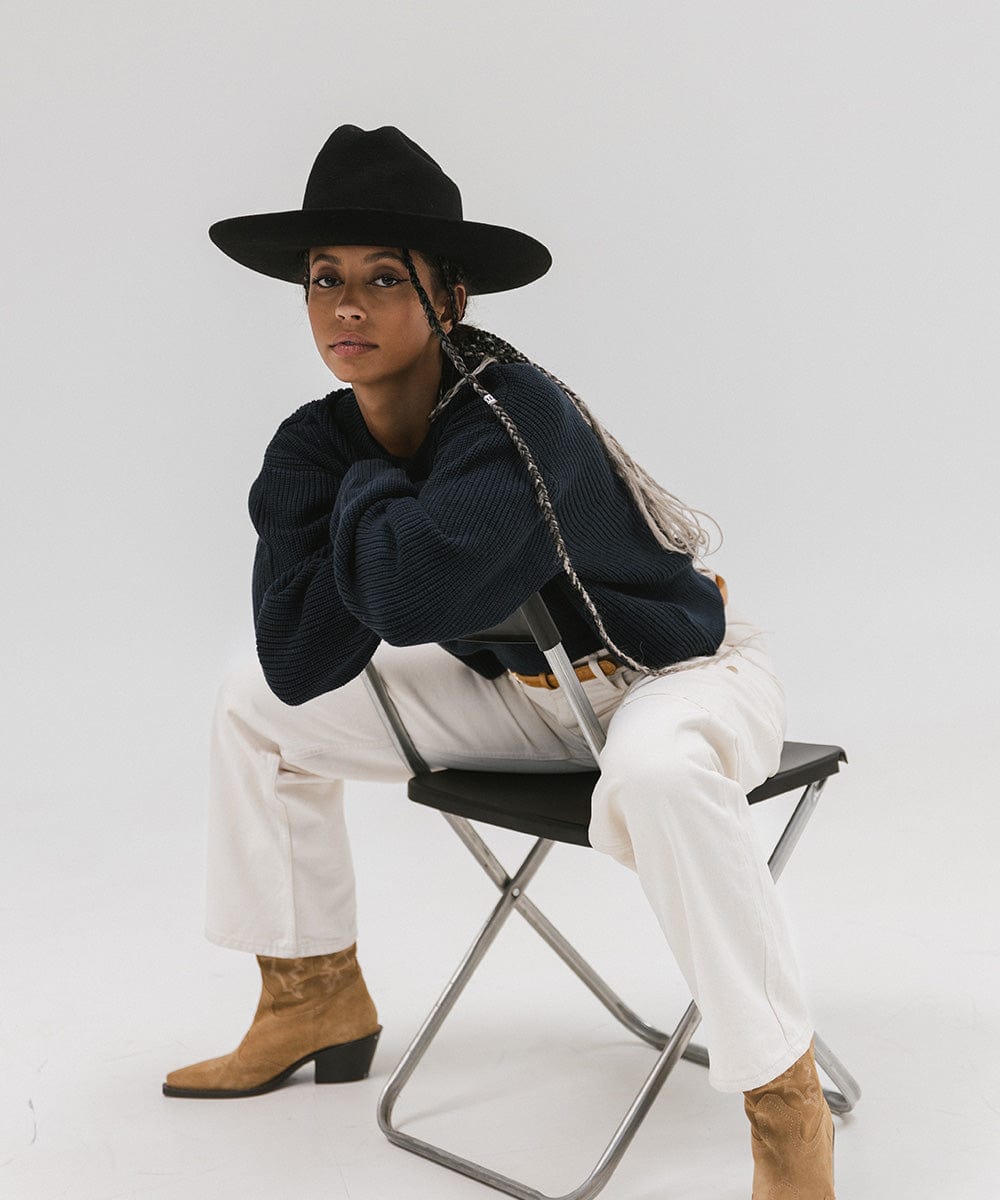 Woman wearing a black Western hat with a classic cattleman crown, a dark blue sweatshirt, white pants, and brown boots, sitting on a chair  against a plain background  #color_black