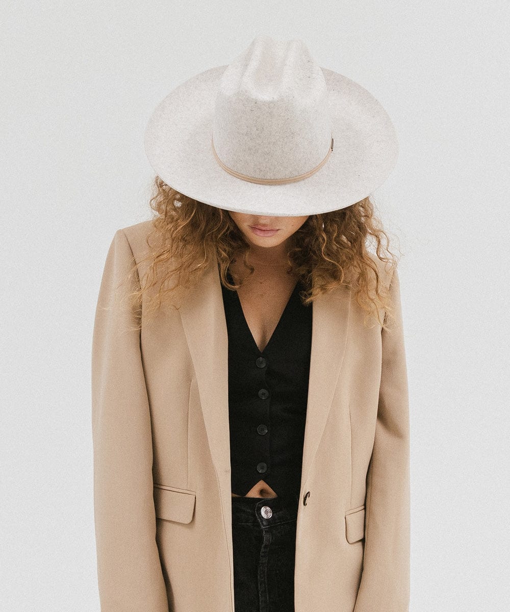 Woman wearing a mix grey colored Western hat with a classic cattleman crown and a hat band, and a beige coat with a black top, looking down against a plain background  #color_mix grey