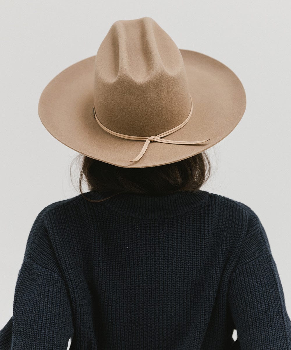 Person wearing a tan Western hat with a classic cattleman crown and a hat band, and a dark blue sweatshirt, facing behind against a plain background  #color_tan
