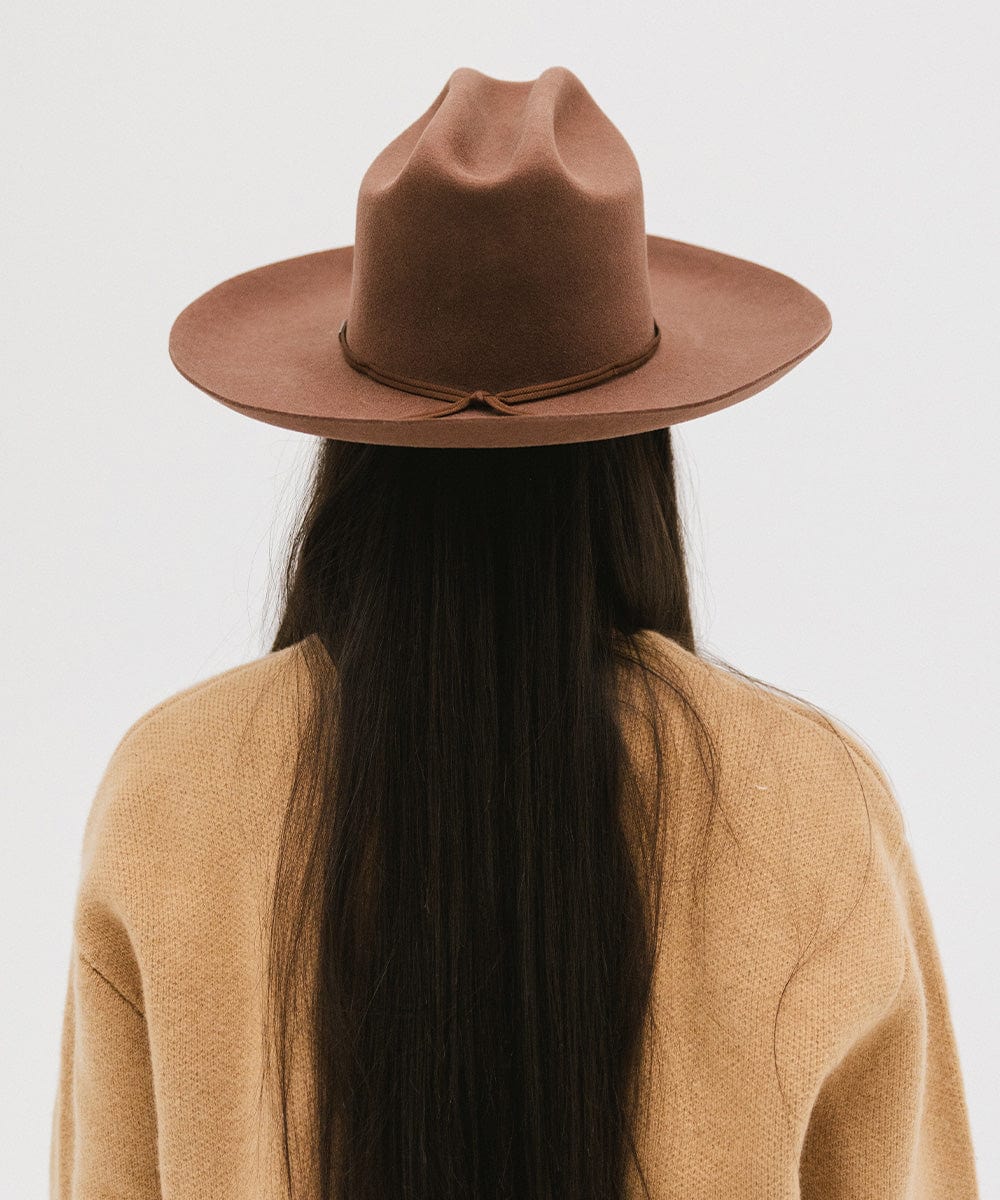 Person with long hair wearing a dark oak colored Western hat with a classic cattleman crown and a hat band, and a brown jacket, facing behind against a plain background  #color_dark oak