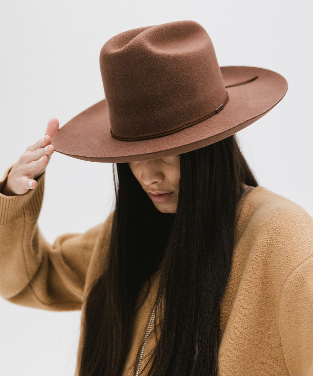 Woman wearing a dark oak colored Western hat with a classic cattleman crown and a hat band, and a brown jacket, looking down against a plain background  #color_dark oak