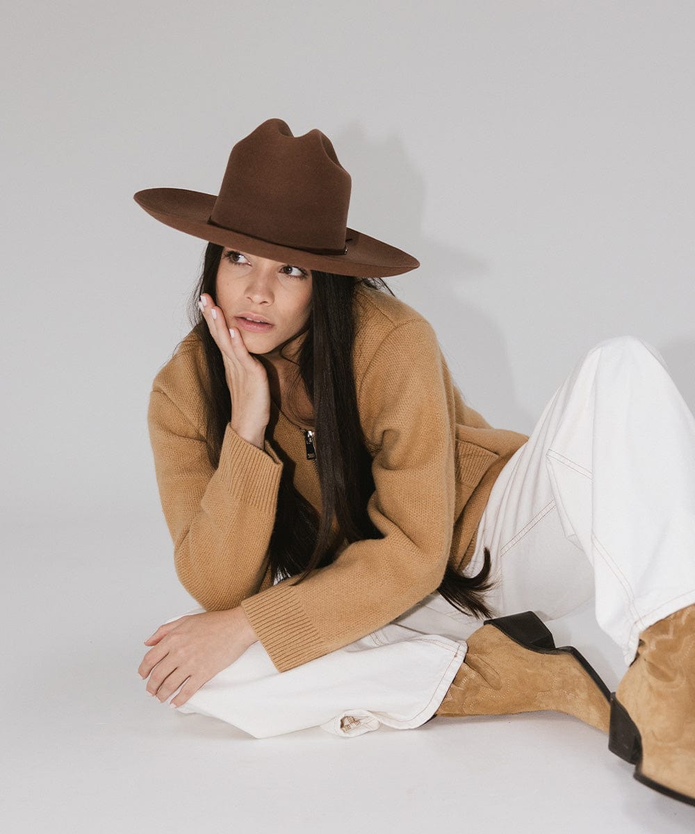 Woman wearing a dark oak colored Western hat with a classic cattleman crown, a brown jacket, white pants and brown boots, sitting against a plain background  #color_dark oak