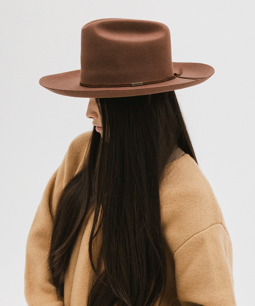 Woman wearing a dark oak colored Western hat with a classic cattleman crown and a hat band, and a brown jacket, facing sideways against a plain background  #color_dark oak