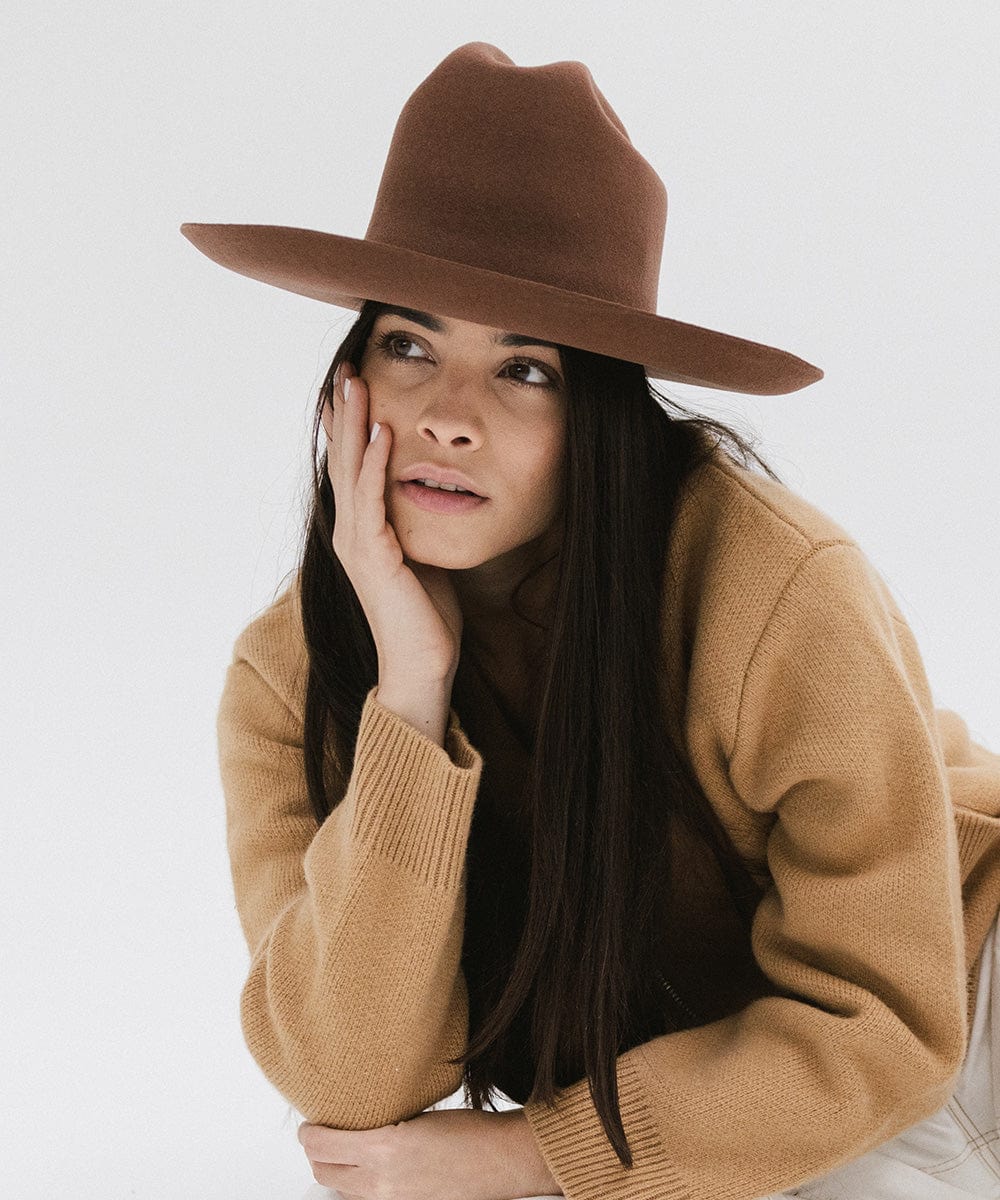 Woman wearing a dark oak colored Western hat with a classic cattleman crown, and a brown jacket, looking sideways against a plain background  #color_dark oak