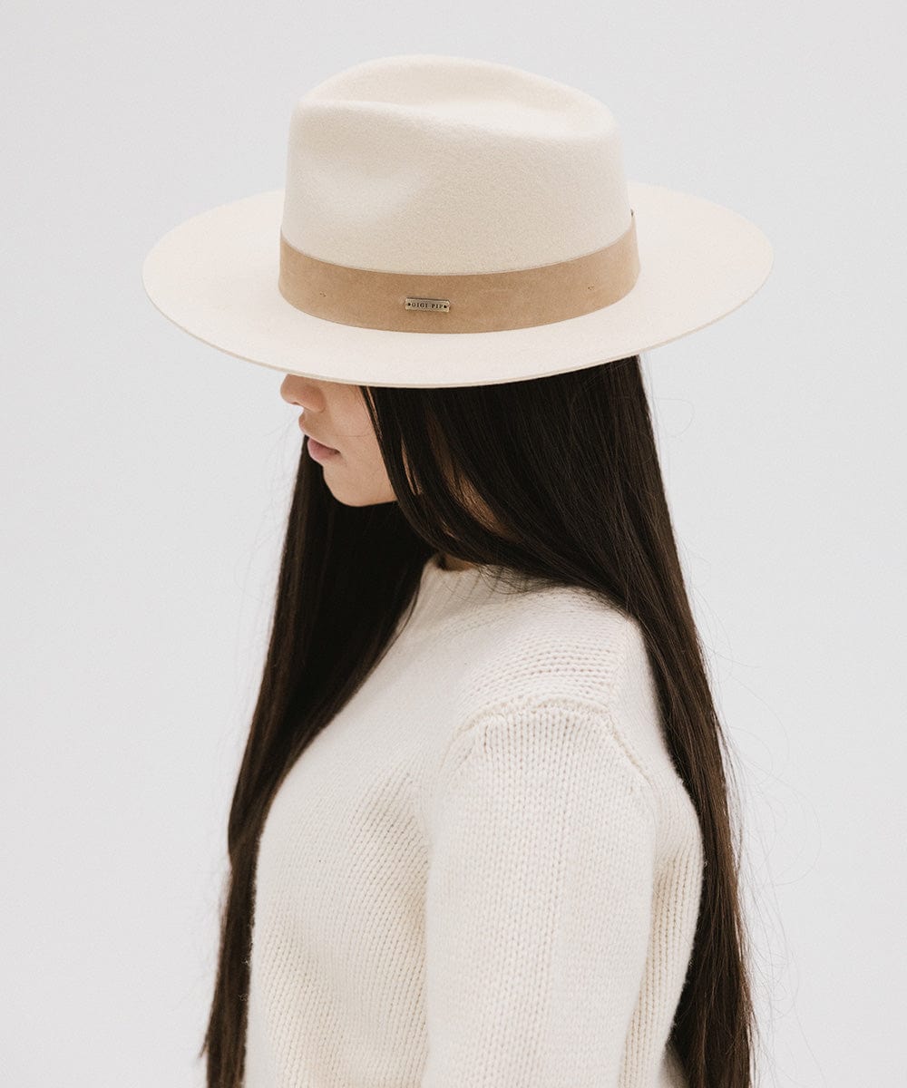 Woman with long hair wearing an off white felt fedora hat with a band and a white sweater, looking sideways against a plain background  #color_off white