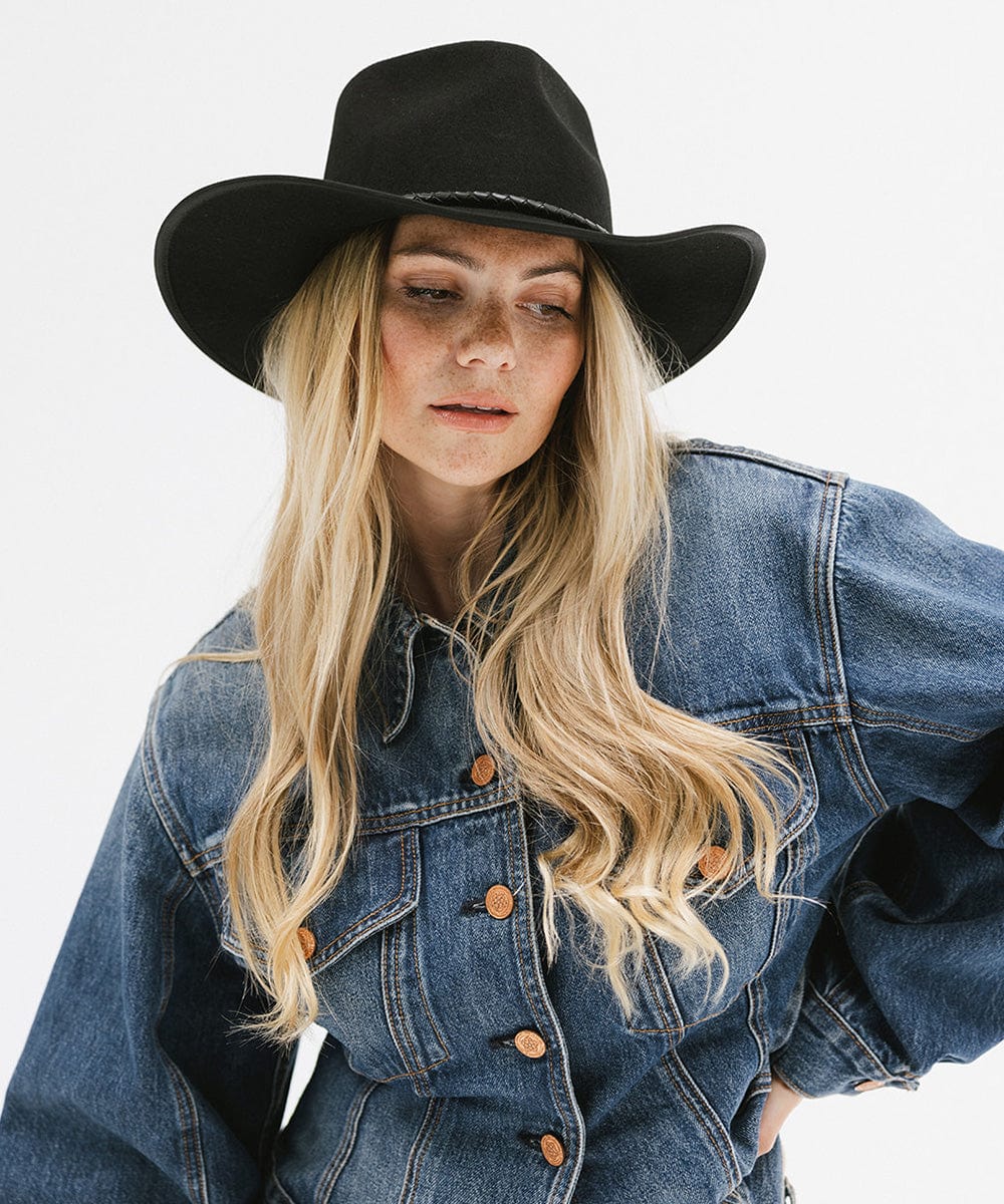 Woman with long blonde hair wearing a black felt western hat and a denim shirt against a white background #color_black