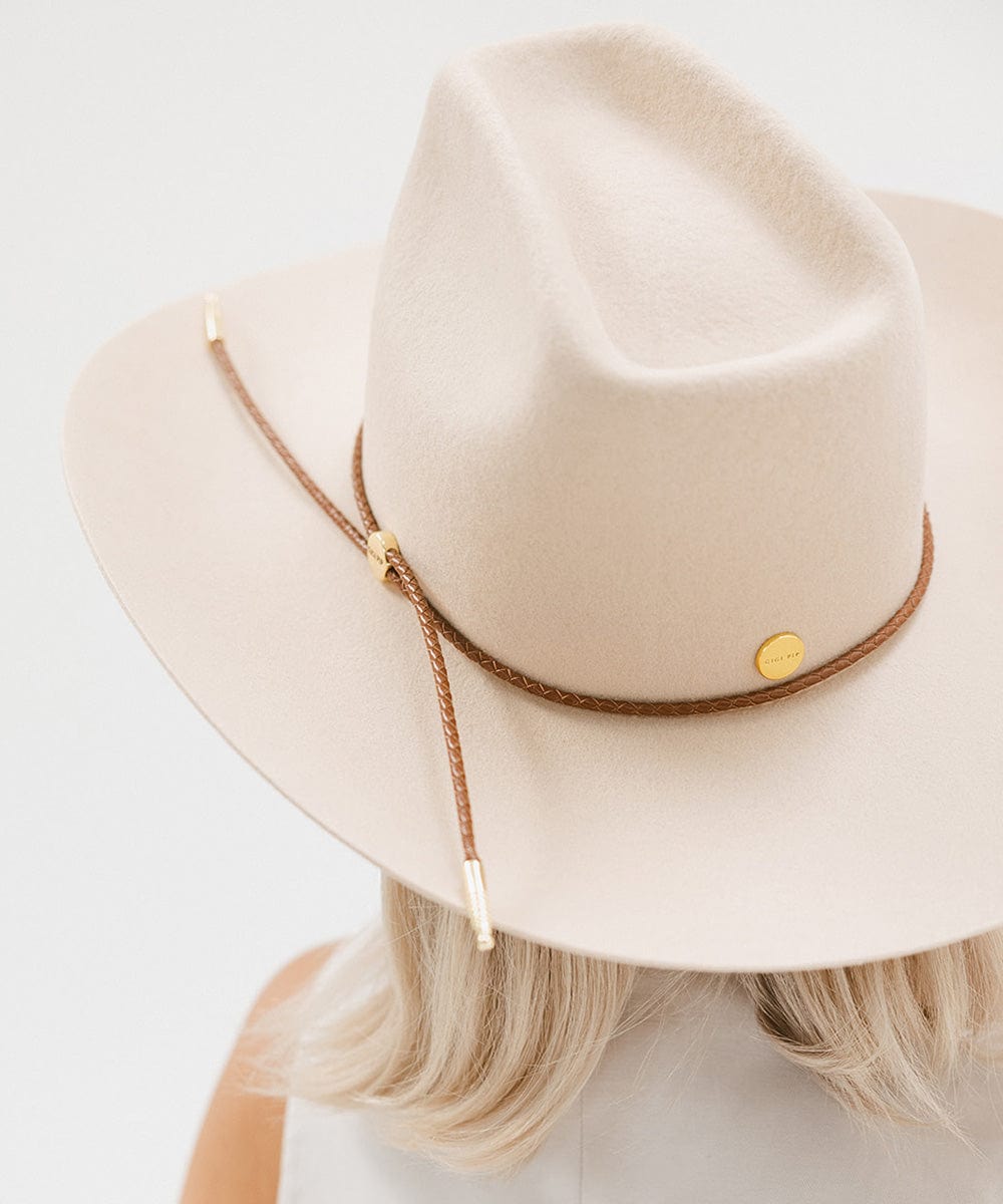 Person wearing a cream cowboy hat with a stiff, traditional wide western brim and a brick top style crown along with a brown hat band against a plain background #color_cream