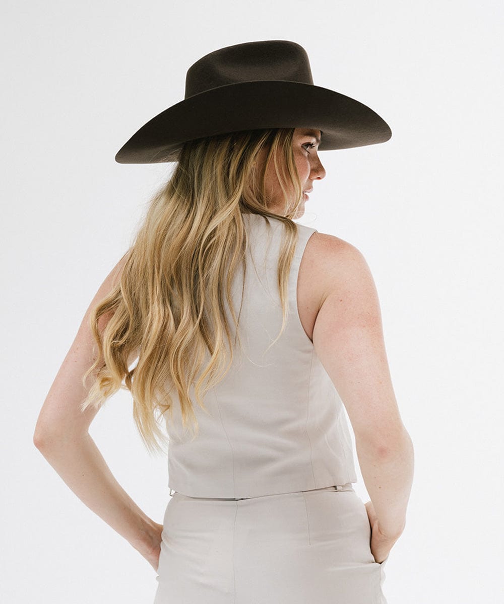 Woman with long hair wearing a dark brown cowboy hat and a white sleeveless jacket, looking sideways against a plain background #color_dark brown