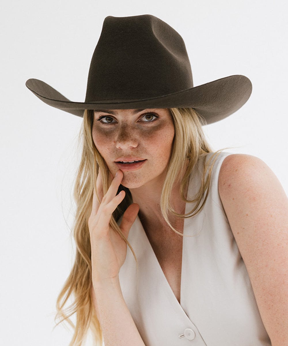 Woman wearing a dark brown cowboy hat and a white sleeveless jacket, posing against a plain background #color_dark brown