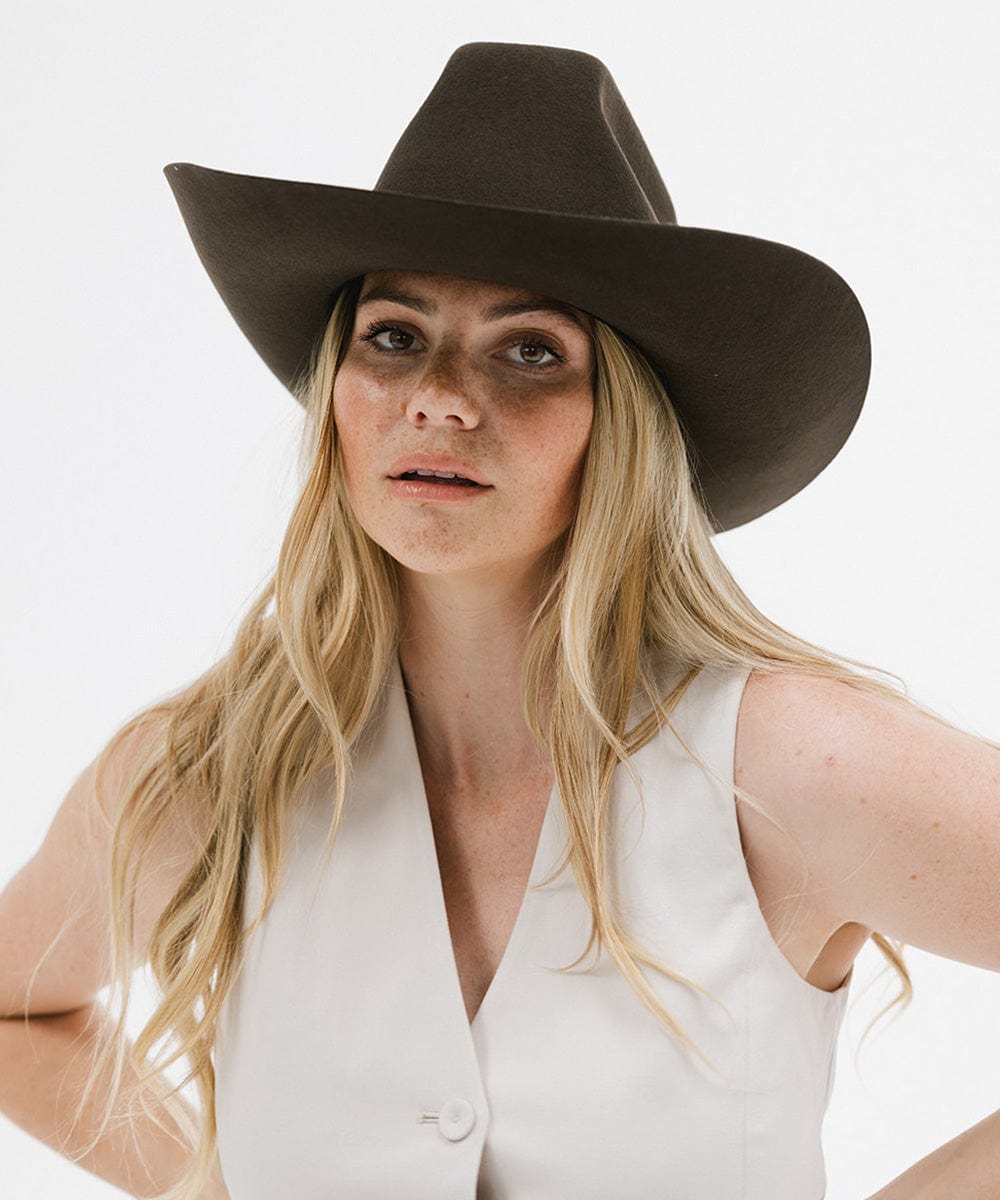 Woman blonde hair wearing a dark brown cowboy hat and a white sleeveless jacket against a plain background #color_dark brown