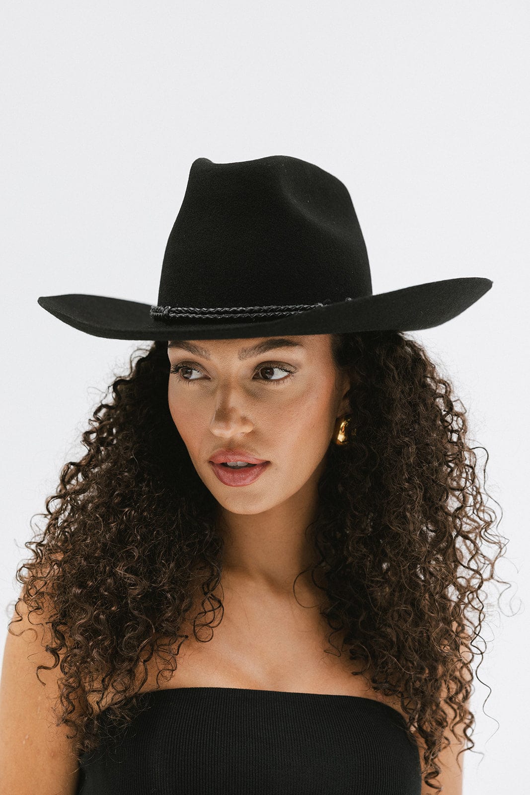 Woman with curly hair wearing a black cowboy hat with a black hat band and a black top, looking sideways against a plain background #color_black