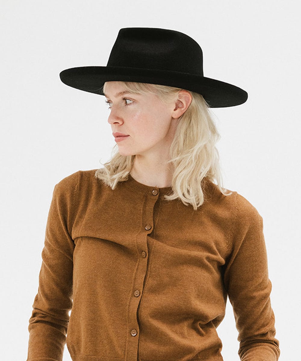 Woman wearing a black felt fedora hat with a hat band and a brown sweater against a plain background #color_black