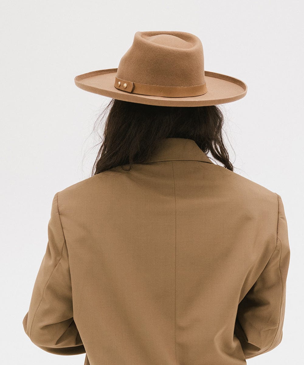 Person wearing a tan felt fedora hat with a hat band and a brown coat against a plain background #color_tan