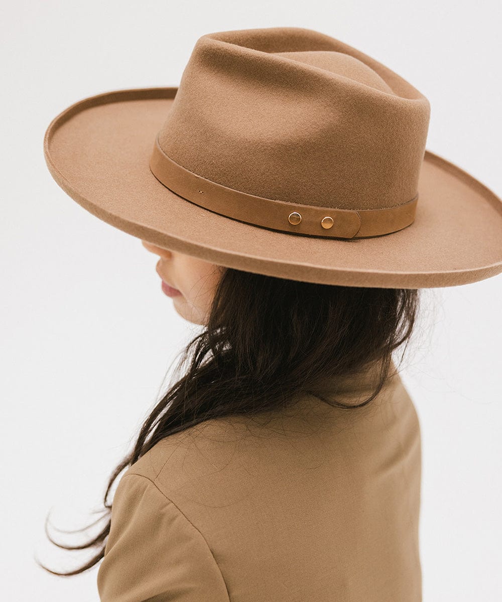 Woman wearing a tan felt fedora hat with a hat band and a brown coat, facing behind against a plain background #color_tan