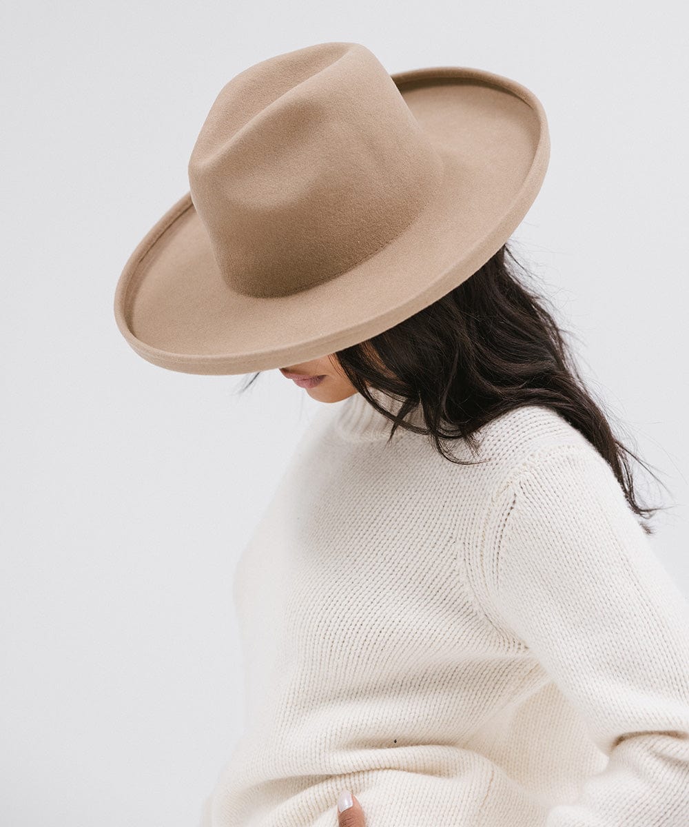 Woman wearing a tan pencil brim felt fedora hat and a white sweatshirt, looking down against a white background #color_tan