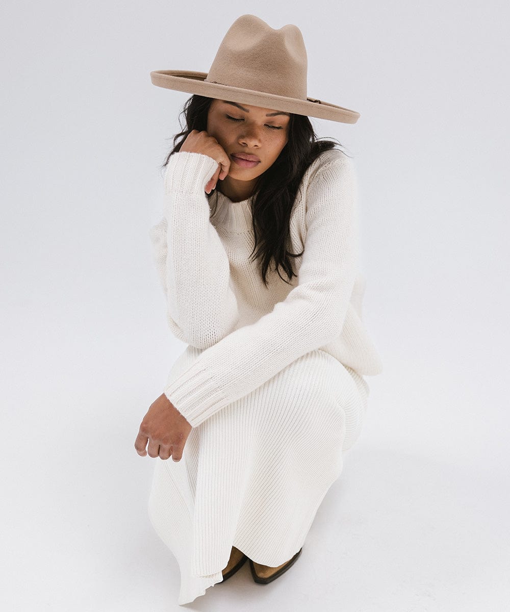 Woman wearing a tan pencil brim felt fedora hat and a white outfit, sitting against a white background #color_tan