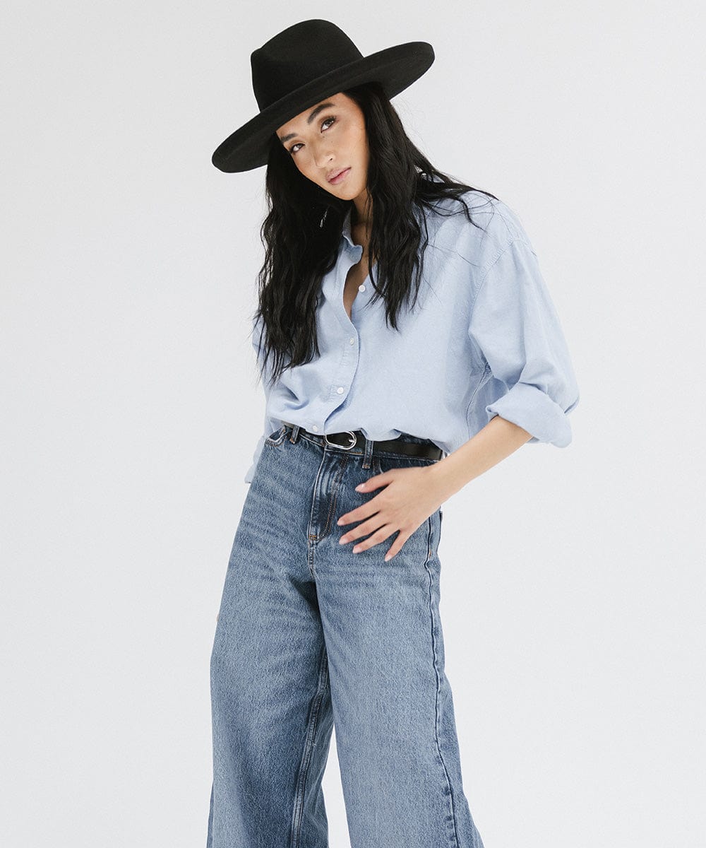 Woman wearing a black pencil brim felt fedora hat, a blue shirt and blue jeans against a white background #color_black