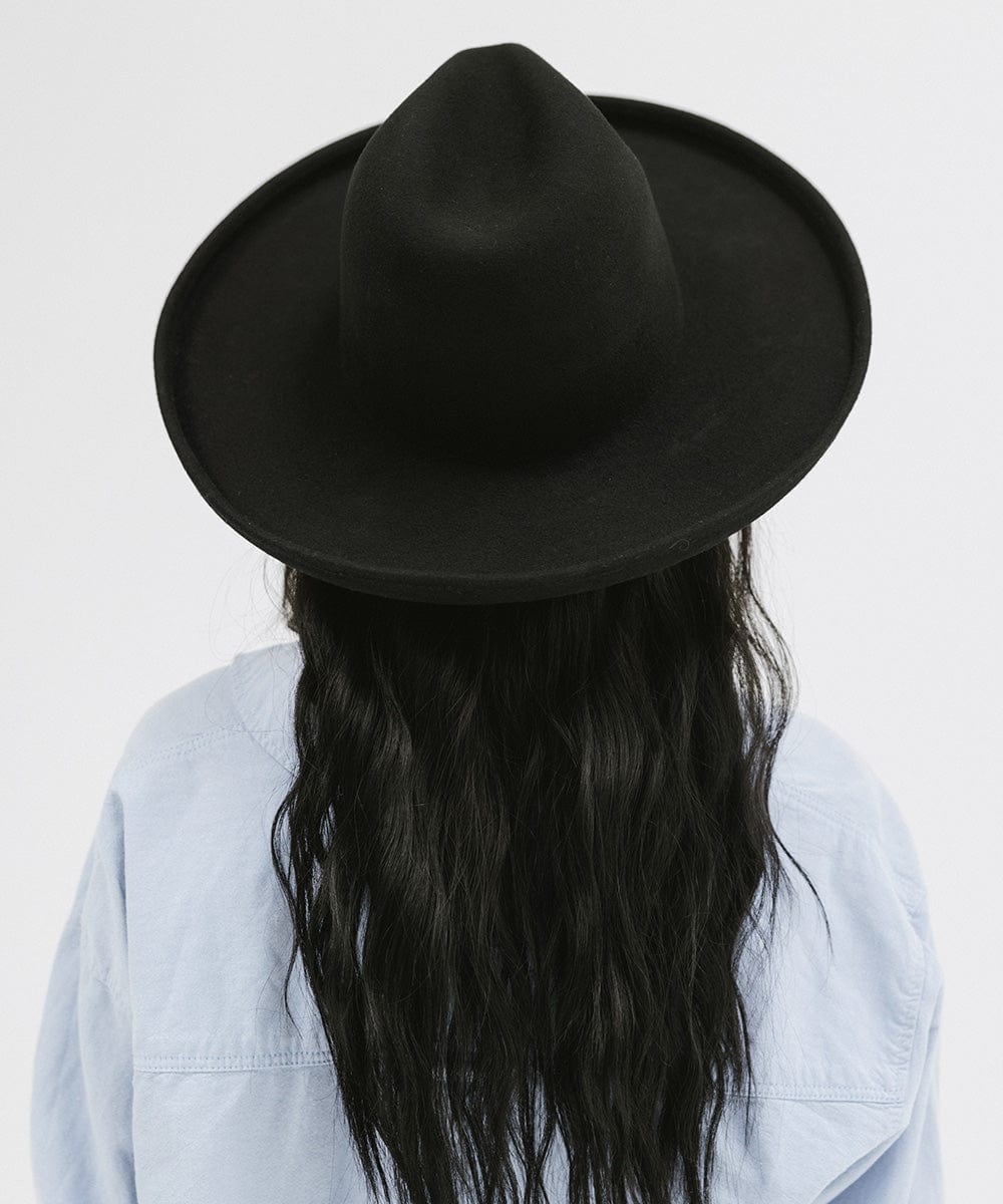 Woman wearing a black pencil brim felt fedora hat and a blue shirt, facing behind against a white background #color_black
