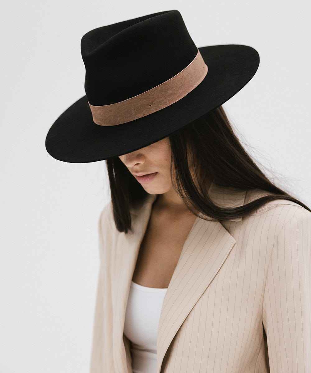 Woman wearing a black fedora hat with a brown band and a cream blazer, looking down against a white  background #color_black-brown