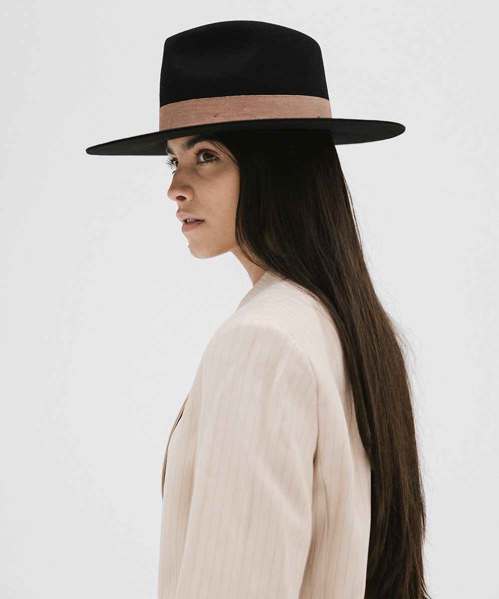 Woman with long hair wearing a black fedora hat with a brown band and a cream blazer, looking sideways against a white background #color_black-brown