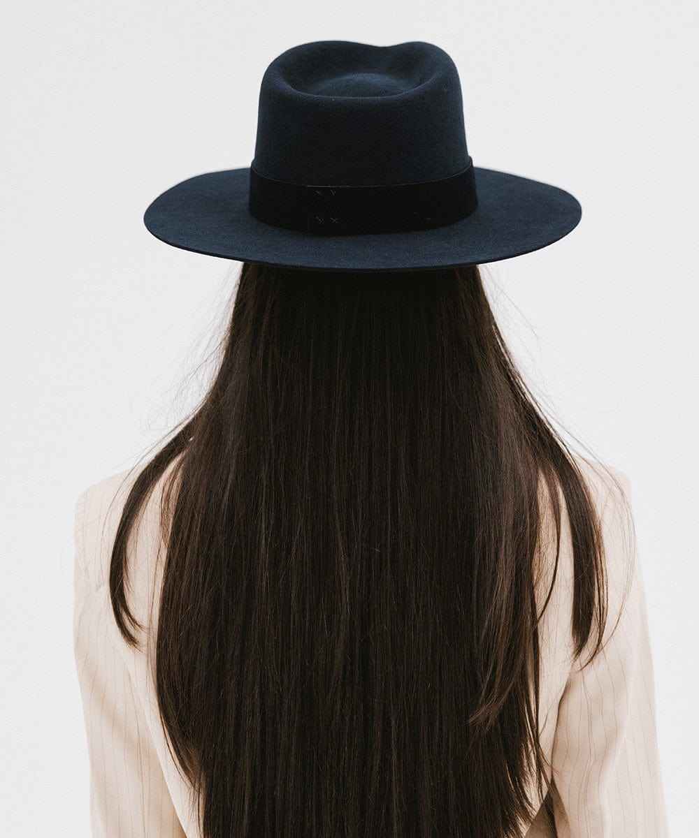 Woman with long hair wearing a navy fedora hat with a band facing behind against a plain background #color_navy