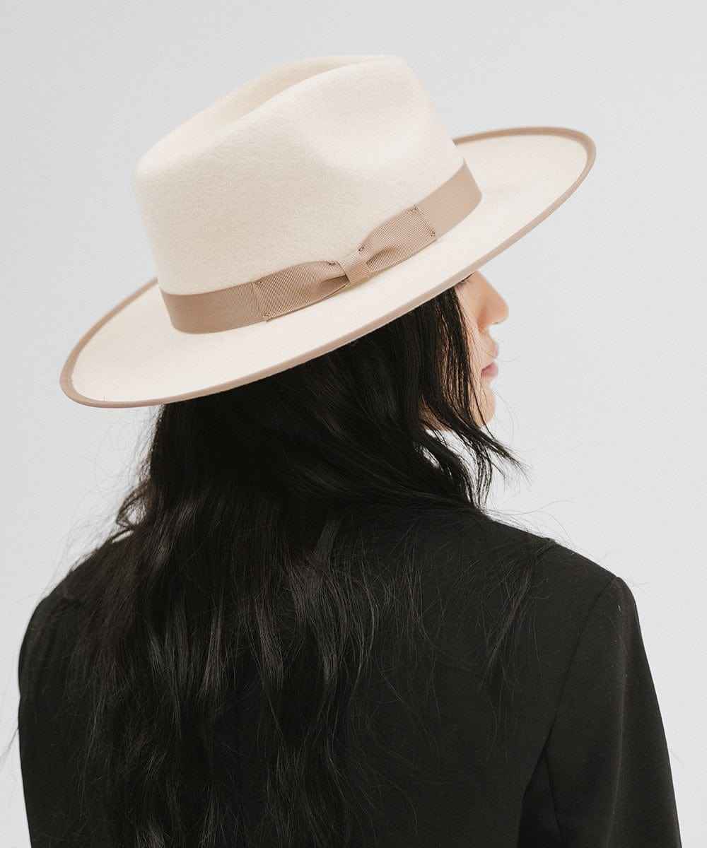 Woman wearing a white and taupe colored fedora hat with a taupe band and a black blazer, facing behind against a plain background #color_white-taupe