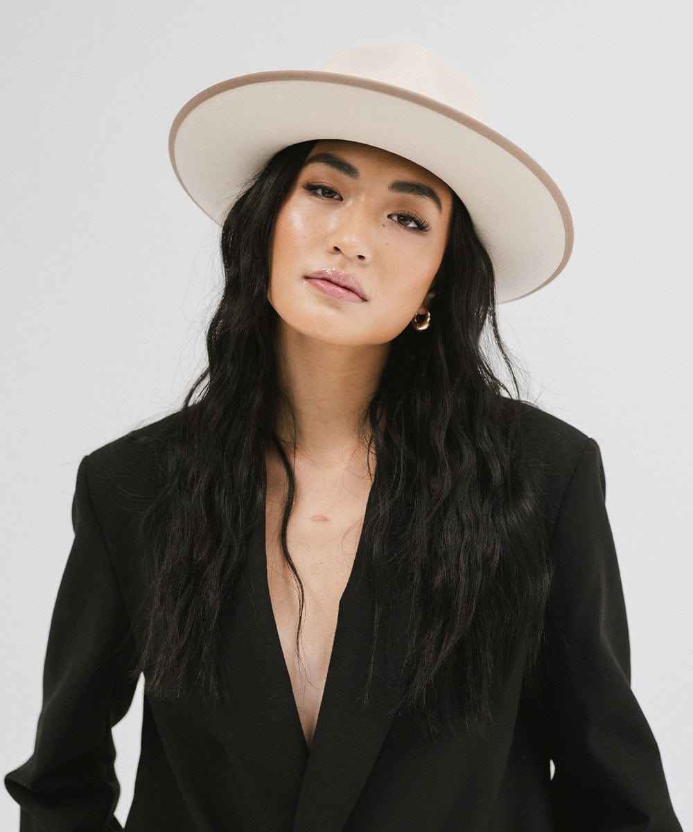Woman wearing a white and taupe colored fedora hat with a taupe band and a black blazer against a plain background #color_white-taupe