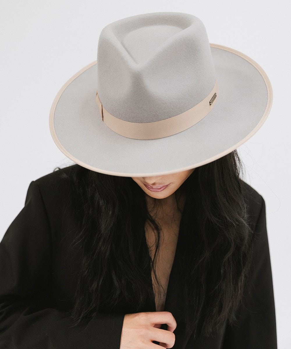 Woman wearing a light grey and tan colored fedora hat with a tan band and a black blazer against a plain background #color_light grey-tan