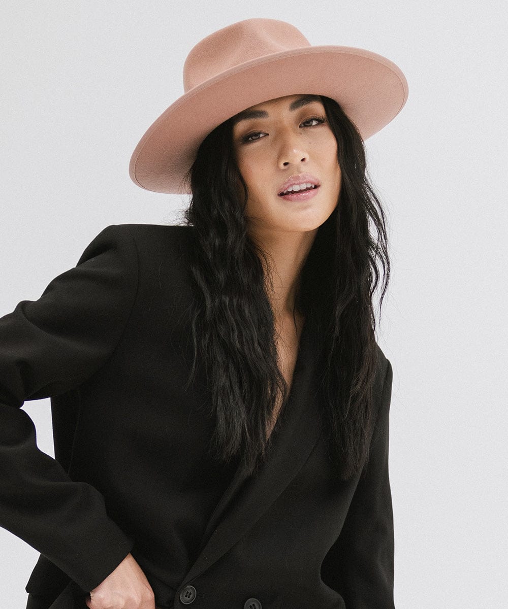 Woman wearing a dusty pink colored fedora hat with a band and a black blazer against a plain background #color_dusty pink