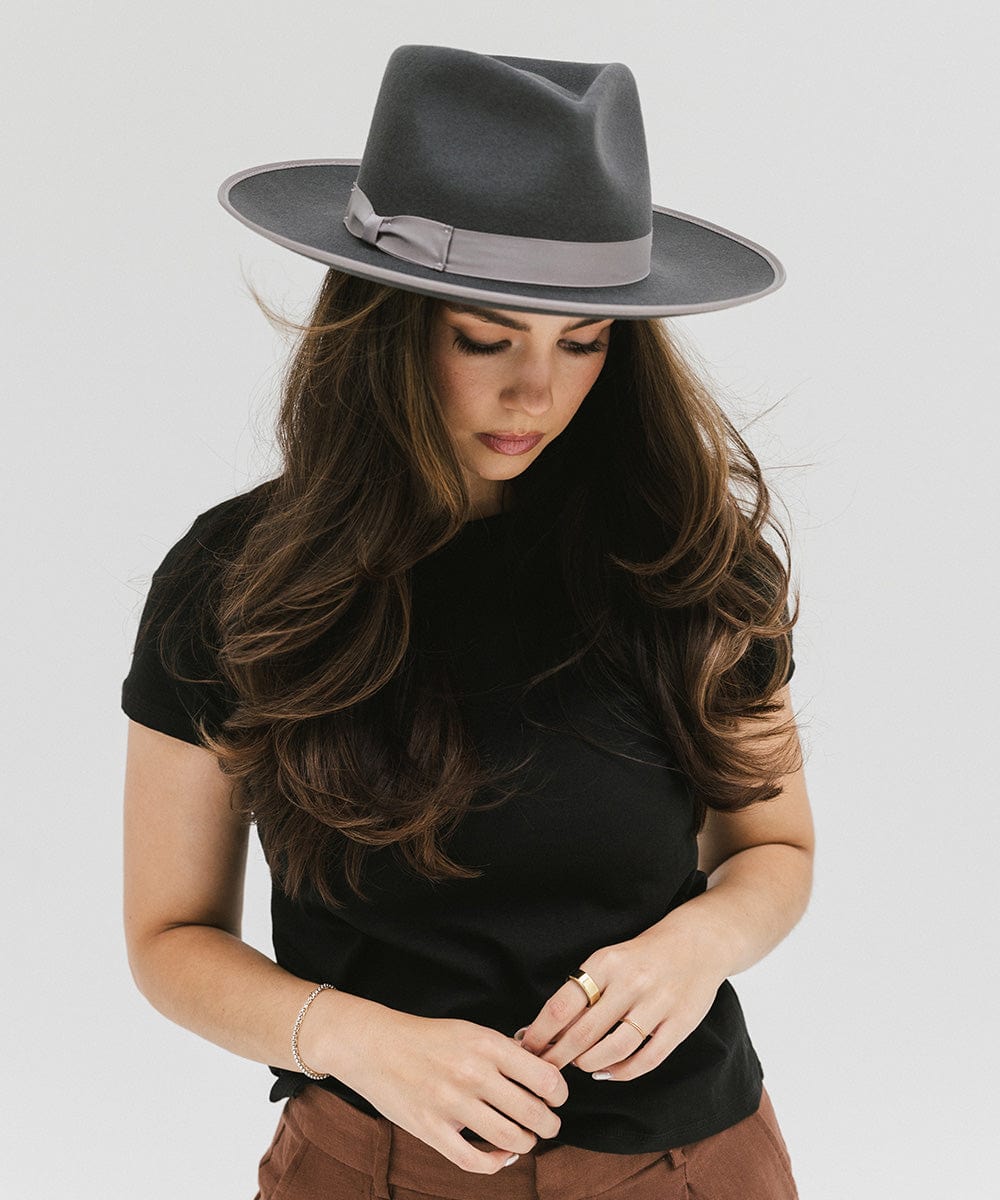 Woman wearing a dark grey fedora hat with a band and a black t-shirt, looking down against a plain background #color_dark grey