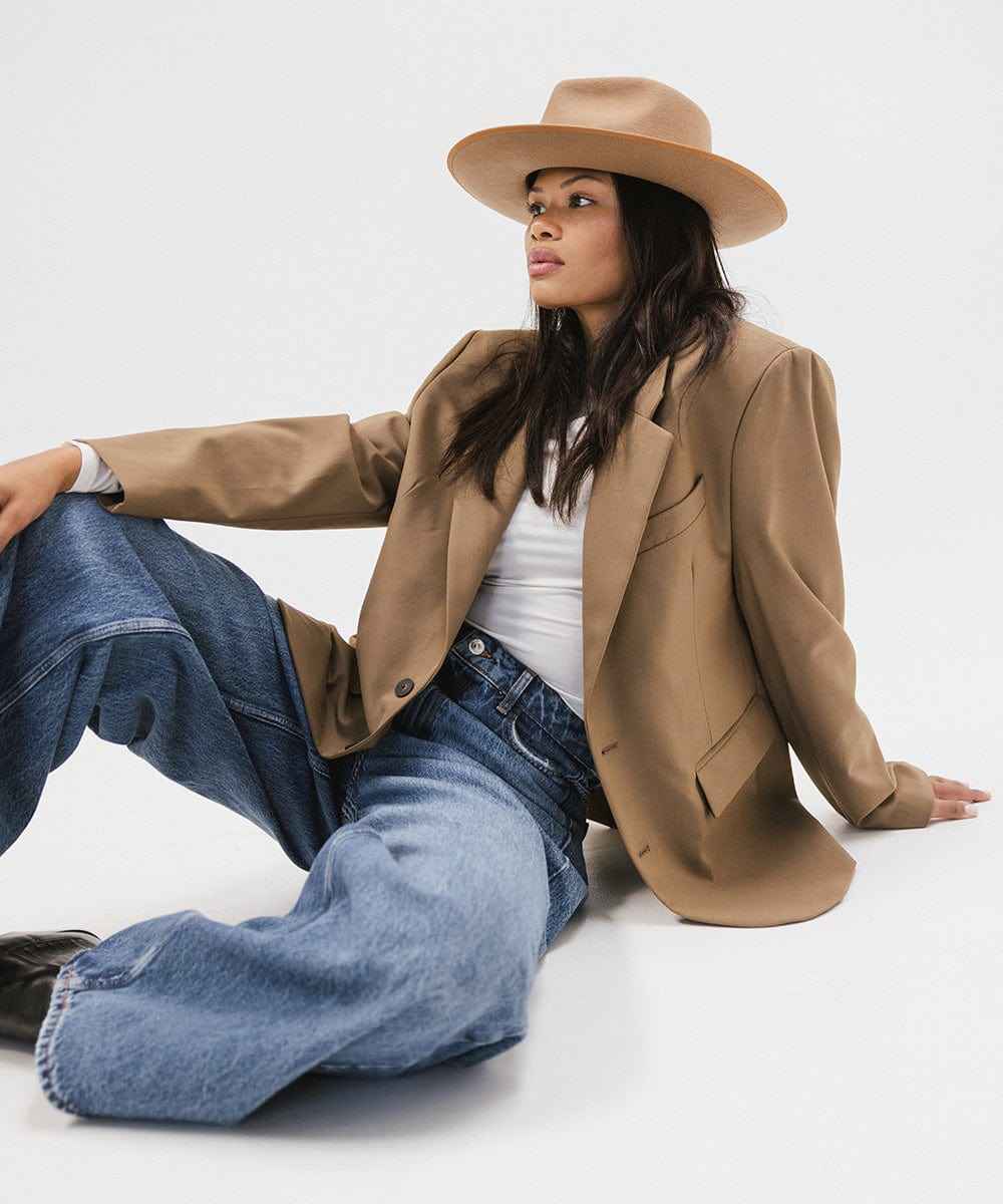 Woman wearing a brown hat with a band, a white top with a brown blazer and blue jeans, sitting and looking sideways against a plain background #color_brown