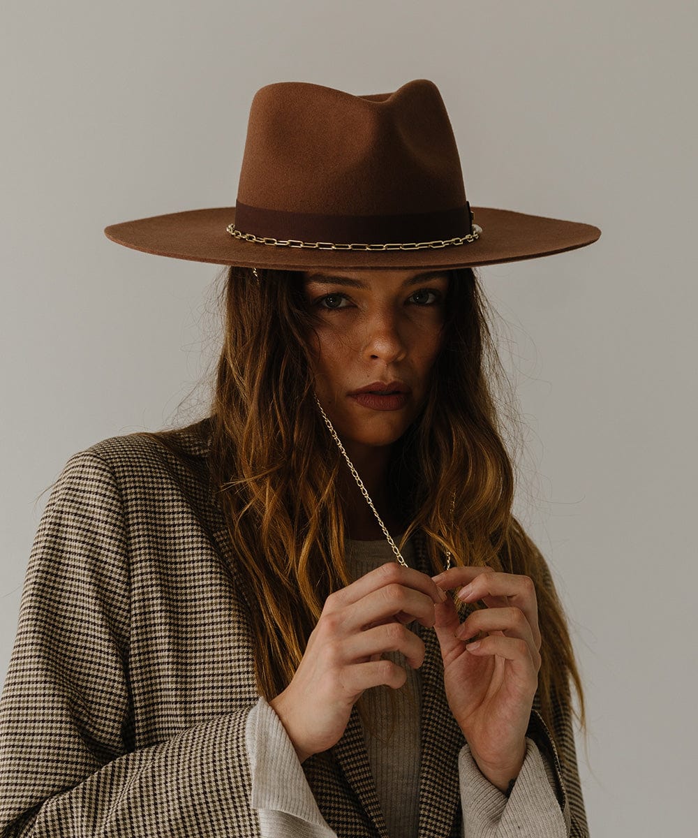 Woman wearing a dark oak colored wide brim fedora hat with a gold chain detail around the base of the crown and a coat, posing against a plain background  #color_dark oak