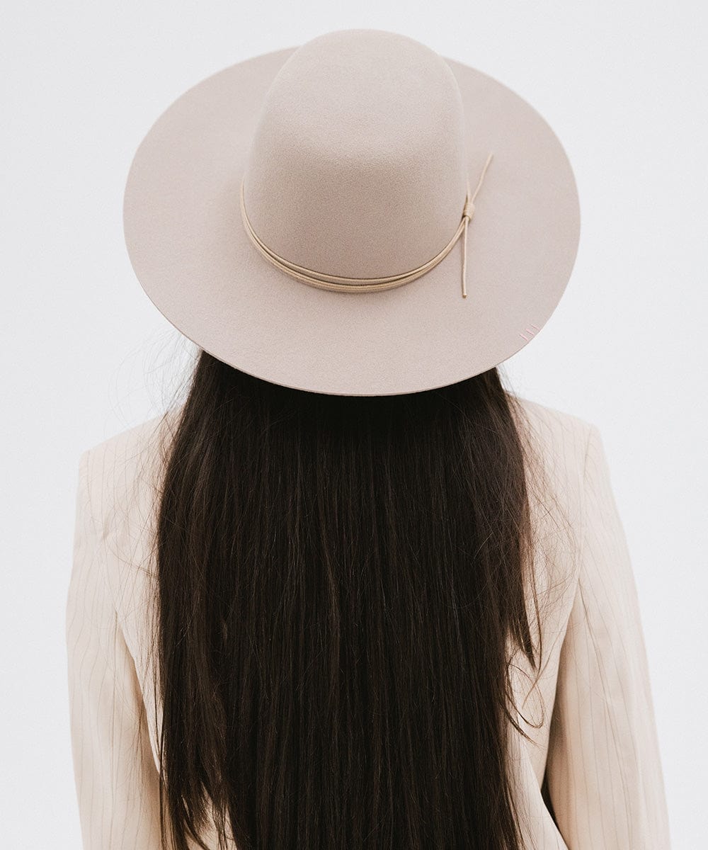 Woman with long hair wearing a sand colored open crown hat with a hat band and a cream blazer against a plain background #color_sand