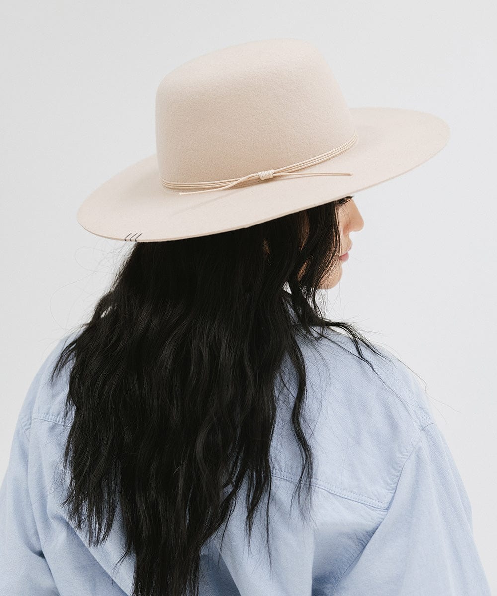 Woman with long black hair wearing a cream open crown hat with a hat band and a blue shirt, looking sideways against a plain background  #color_cream