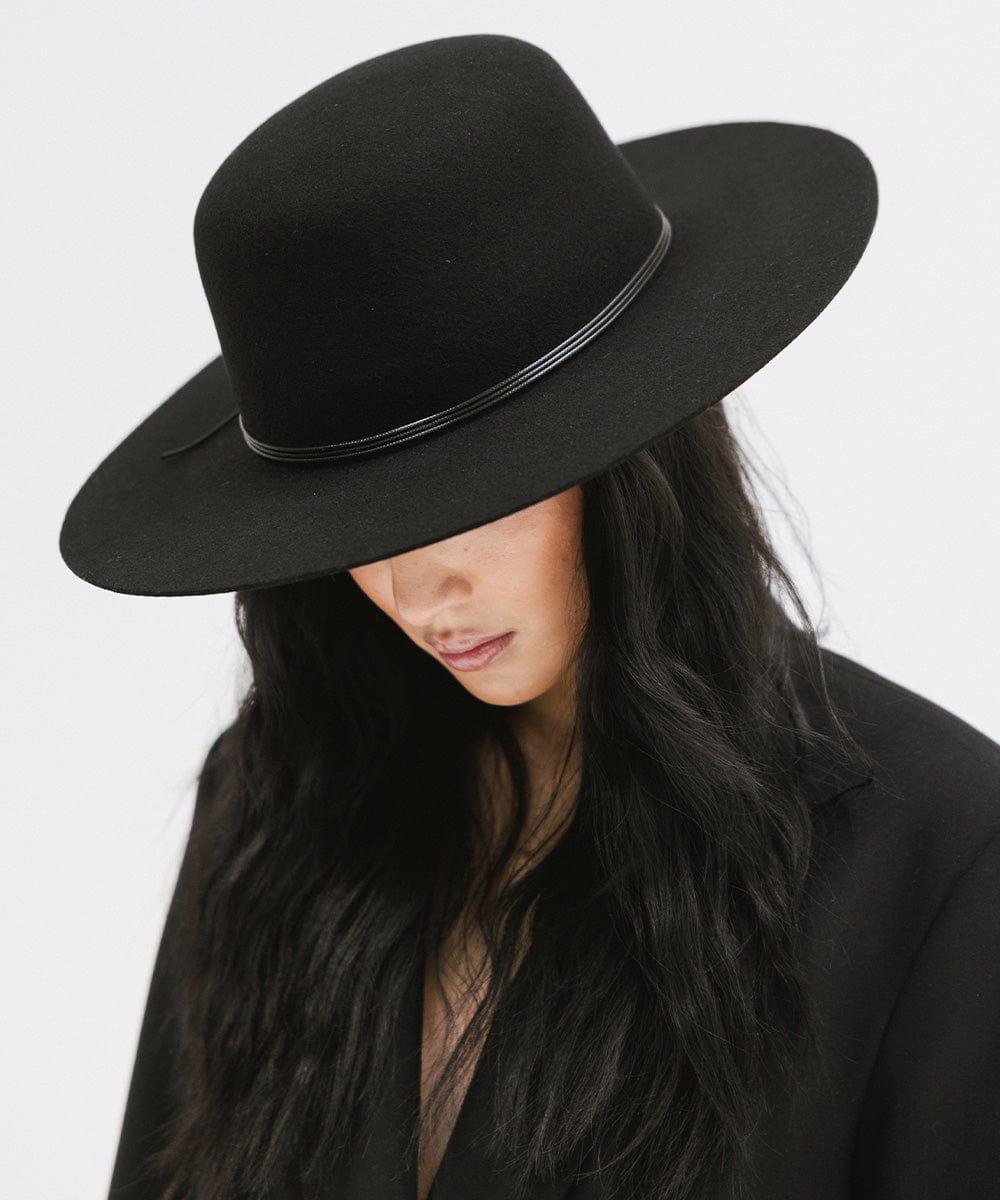 Woman wearing a black open crown hat with a hat band and a black blazer, posing against a plain background #color_black