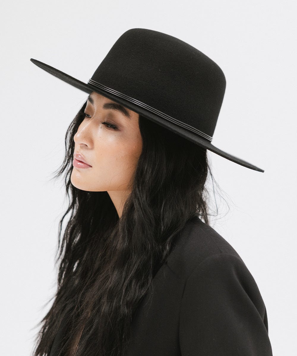 Woman wearing a black open crown hat with a hat band and a black blazer, facing sideways against a plain background #color_black