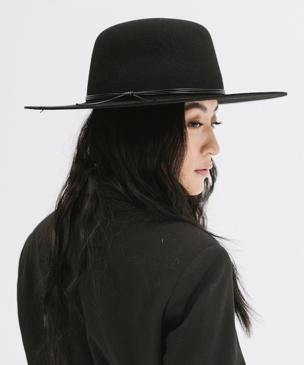 Woman wearing a black open crown hat with a hat band and a black blazer, posing sideways against a plain background #color_black