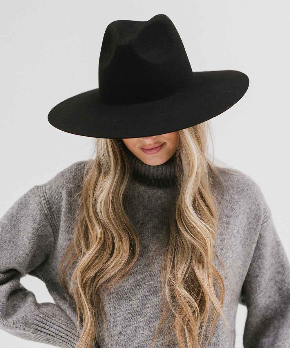 Woman wearing a black wide brim fedora hat and a grey sweater, looking down against a plain background #color_black